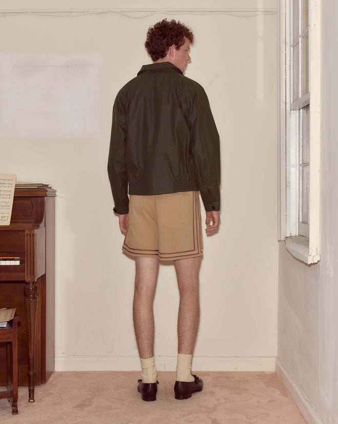 A man wearing the Waxed Coach's Jacket in green, paired with shorts and loafers, stands facing a corner of a beige room near a piano.