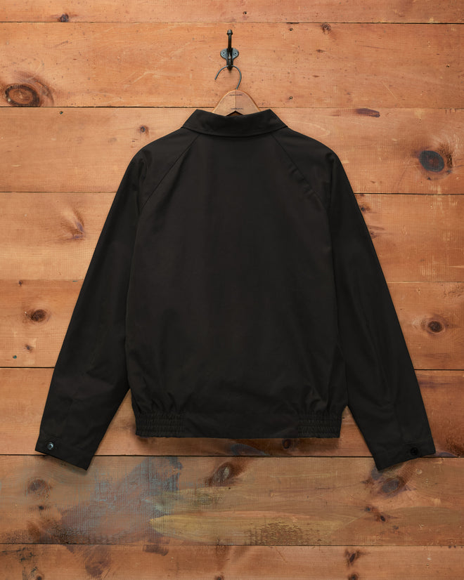 The Waxed Coach’s Jacket - Black is displayed hanging on a wall with wooden planks, shown from the back with its collar and sleeves visible.