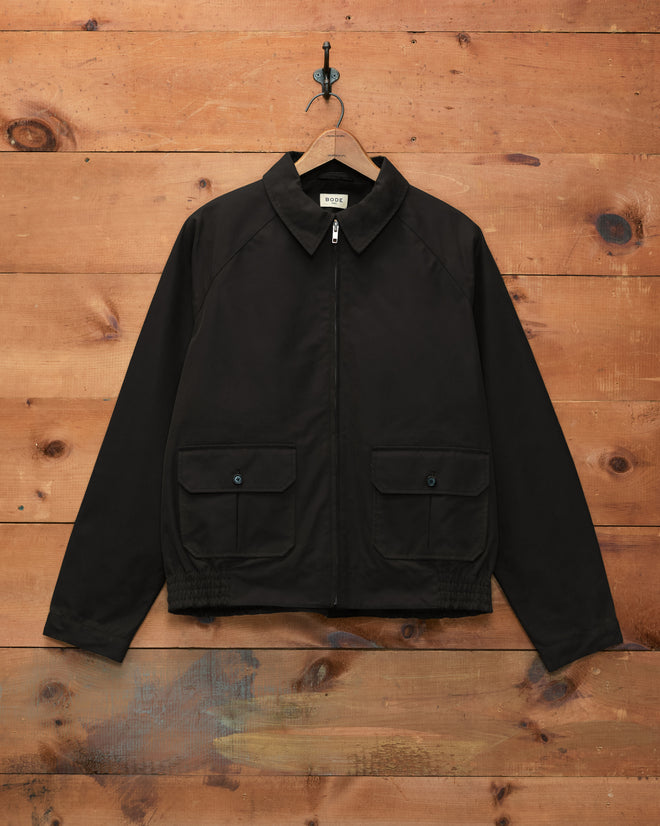 Waxed Coach’s Jacket - Black, featuring a zip-up front and two front pockets, displayed against a wooden wall background.