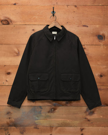 Waxed Coach’s Jacket - Black, featuring a zip-up front and two front pockets, displayed against a wooden wall background.