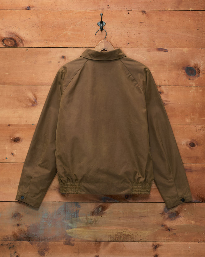 A Waxed Coach’s Jacket in Caramel hangs on a hook against a wooden wall, displayed with its back facing forward—a classic piece of outerwear that exudes timeless style.