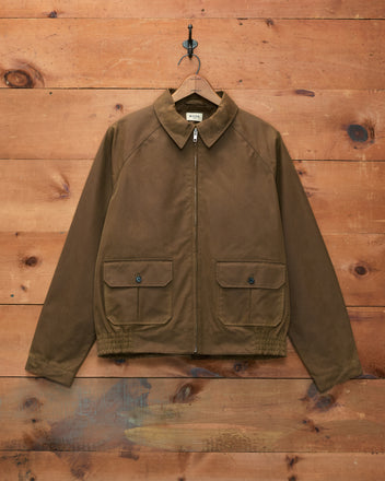 The Waxed Coach’s Jacket in Caramel, featuring two front pockets and crafted from waxed cotton, hangs on a wooden wall—this timeless outerwear piece seamlessly blends classic style with practical design.