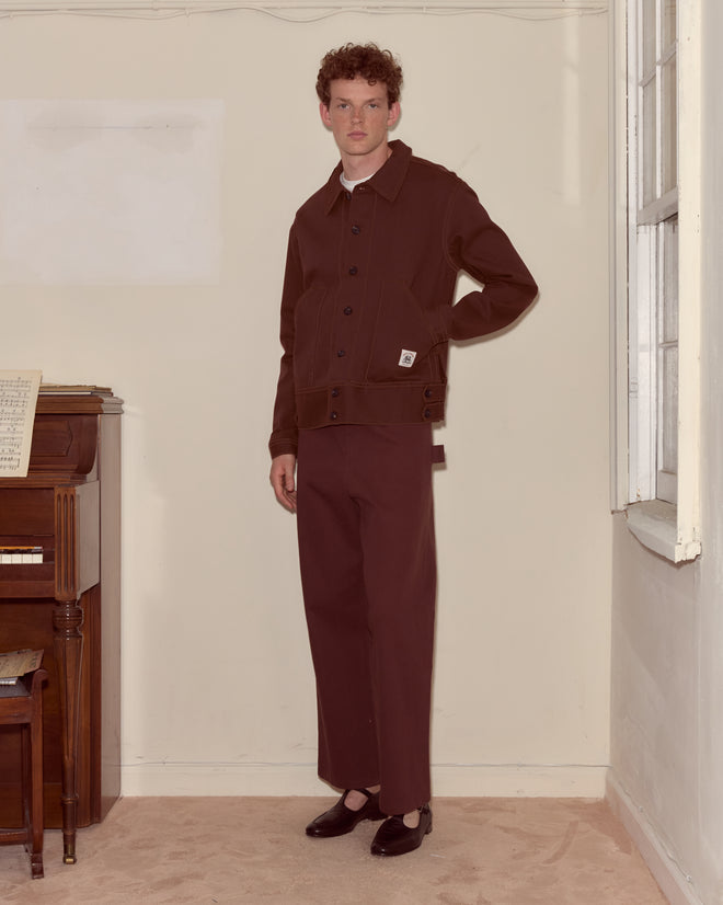 Wearing the Knolly Brook Side Tab Jacket in brown, a person stands indoors by a piano, looking at the camera.