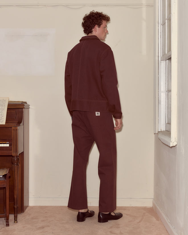 A man in dark clothes and a brown Knolly Brook Side Tab Jacket stands facing a corner near a window, with a piano and sheet music to his left.