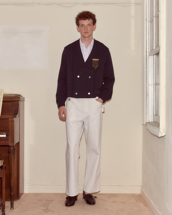 A man in a Double-Breasted Cardigan - Navy and white trousers stands in a minimal room beside a piano and a window.