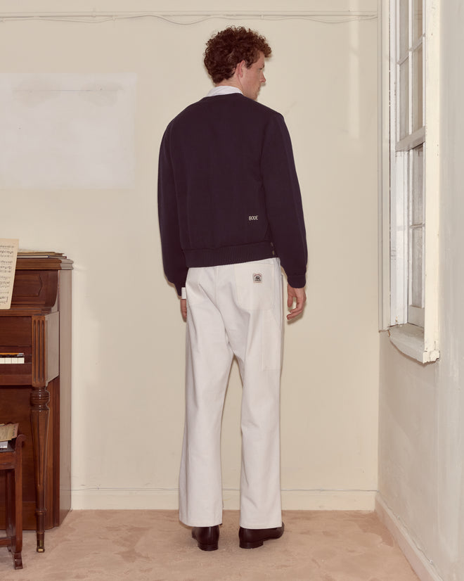 A man stands facing the corner of a room, wearing white pants and a Double-Breasted Cardigan in navy, next to a piano.