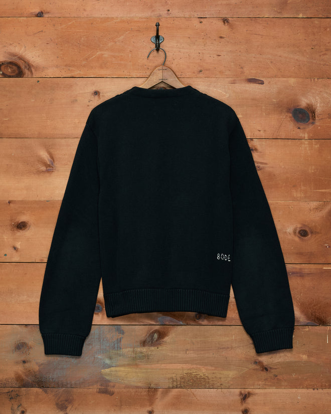 The Double-Breasted Cardigan - Black is displayed on a hanger against a wooden wall, shown from the back with small white text near the hem. Crafted from 100% cotton, this piece seamlessly combines comfort and style in every detail.