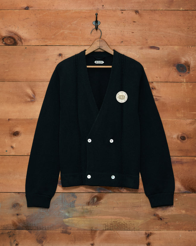 Double-Breasted Cardigan - Black, featuring white buttons and a round hand-embroidered patch, crafted from 100% cotton and displayed against a wooden wall.