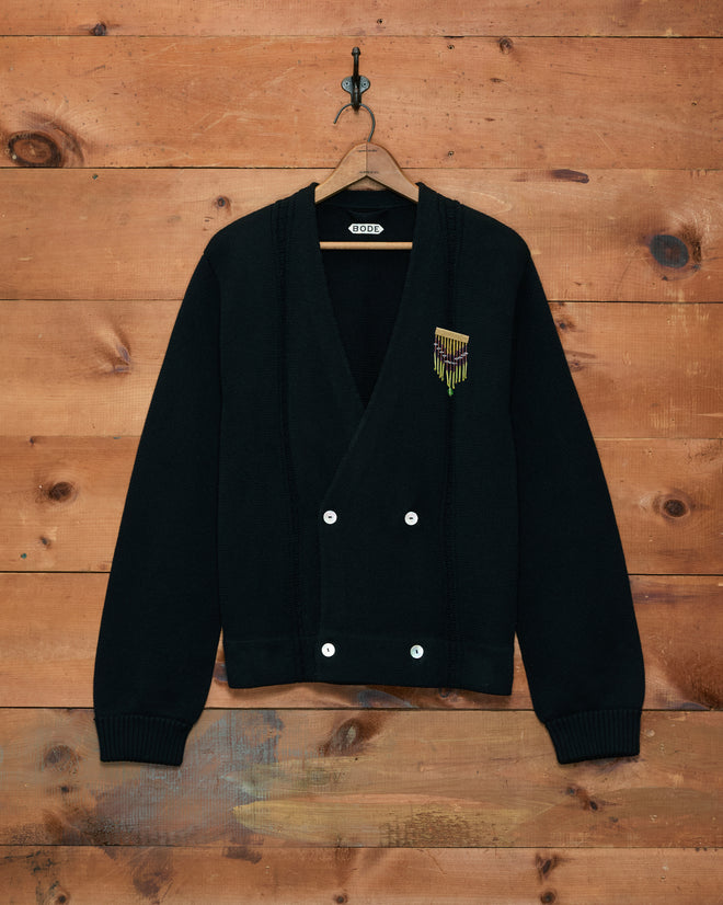 Double-Breasted Cardigan - Black, featuring badge detail and inspired by hand-embroidered Bode knitwear, displayed against a wooden wall background.