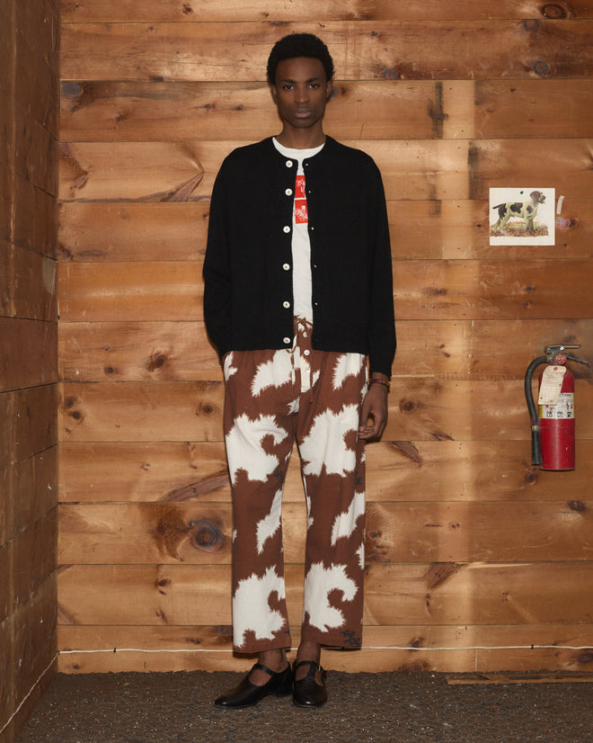Person stands indoors by a wood-paneled wall, wearing the Orchard Cardigan - Black with antique pearl buttons and brown-and-white patterned pants.