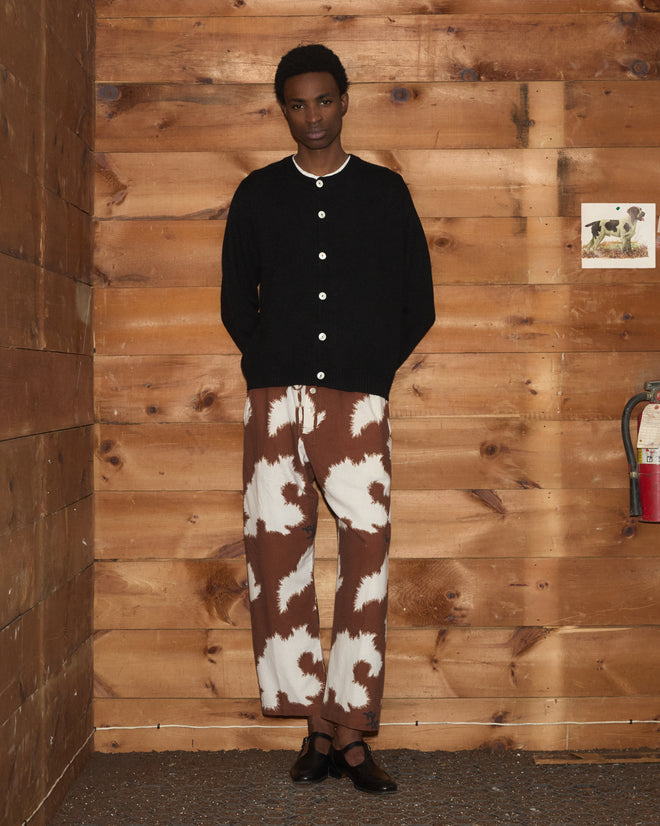 Wearing the Orchard Cardigan - Black, featuring alpaca merino wool and antique pearl buttons, paired with brown-white tie-dye pants, a person stands against a wooden wall with a fire extinguisher nearby.