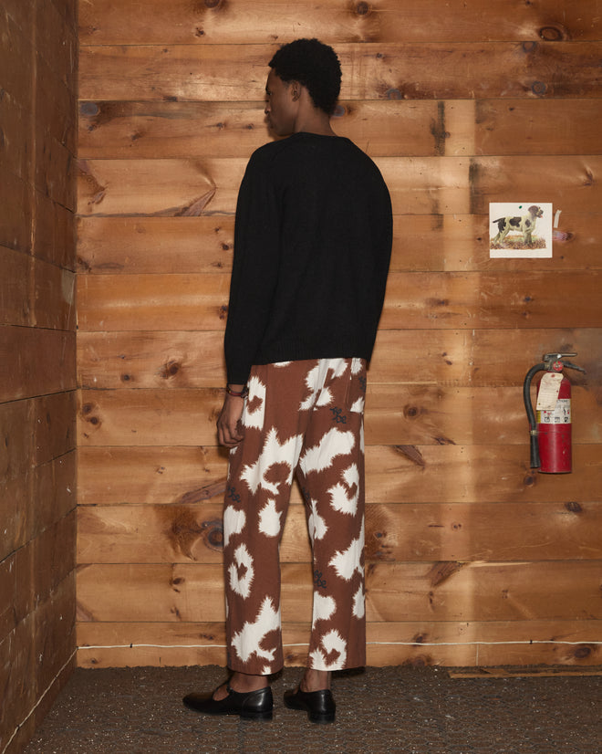 A person stands in a wooden room, dressed in a black shirt and brown patterned pants, facing the wall. Nearby, the Orchard Cardigan - Black—a midcentury children’s sweater crafted from alpaca merino wool and adorned with antique pearl buttons—hangs neatly on a hook, lending timeless character to the serene setting.