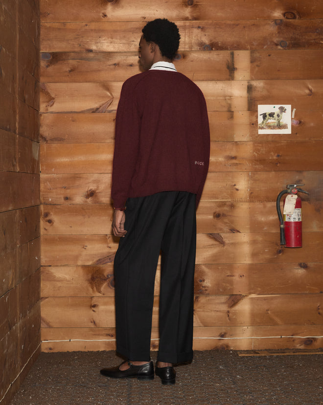 A person wearing the Orchard Cardigan in maroon and black pants stands facing a wooden wall with a fire extinguisher and photo.