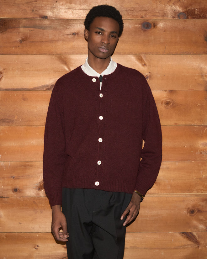 A person wearing the Orchard Cardigan in maroon, inspired by midcentury knitwear, over a white shirt stands against a wooden wall, looking at the camera.