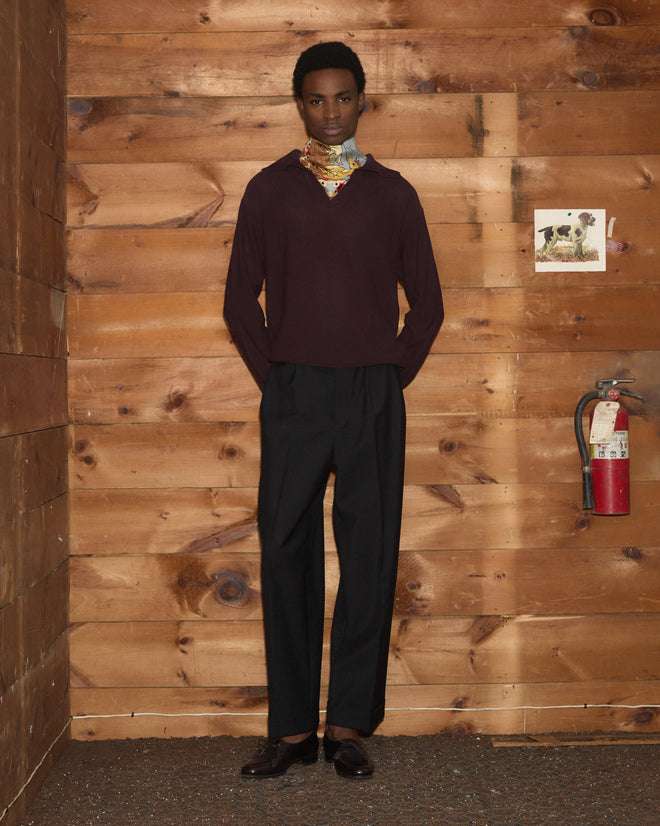 A person wearing the Boxy Long Sleeve Polo - Brown and black pants stands against a wooden wall with a fire extinguisher and a photo.