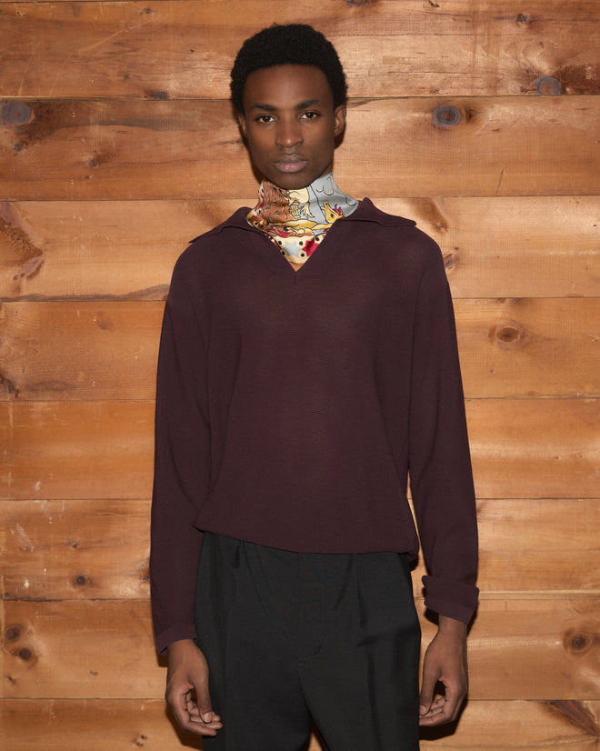 A person wearing the Boxy Long Sleeve Polo in Brown, paired with black pants and a colorful scarf, stands against a wooden wall background.