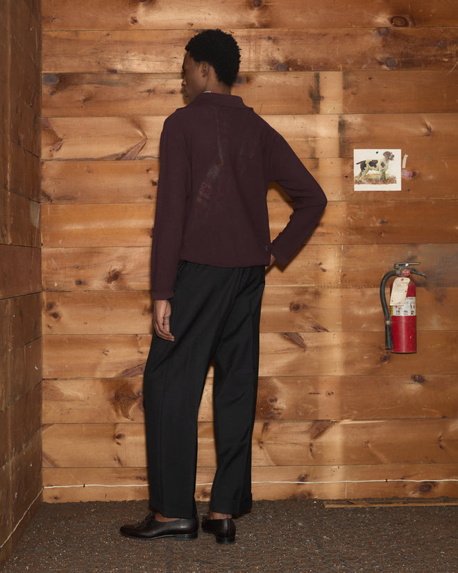 A person wearing the Boxy Long Sleeve Polo in brown and dark pants, standing facing a wooden wall near a fire extinguisher.