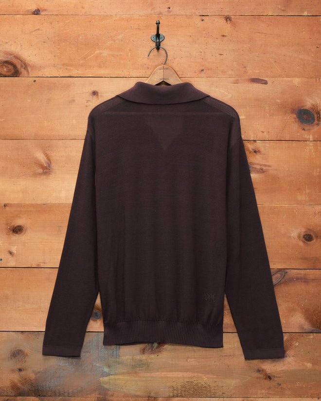 The Boxy Long Sleeve Polo - Brown hangs on a hook against a wooden plank wall, displayed from the back.