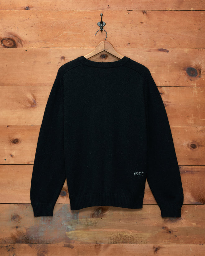 The Orchard Cardigan - Black, crafted from alpaca merino wool and featuring antique pearl buttons, is displayed on a hanger against a wooden wall.