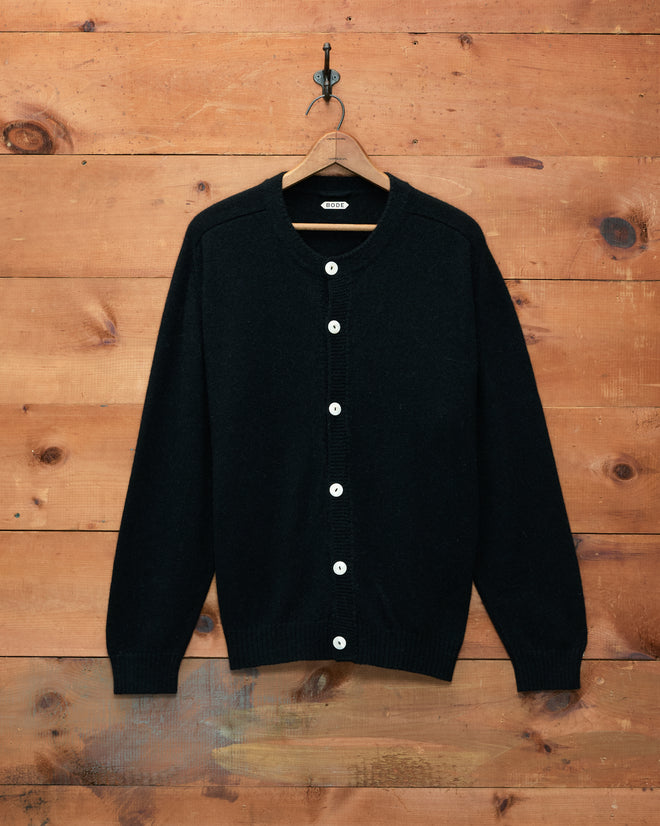 The Orchard Cardigan - Black, displayed on a hanger against a wooden wall, features antique pearl buttons and is crafted from luxuriously soft alpaca merino wool.