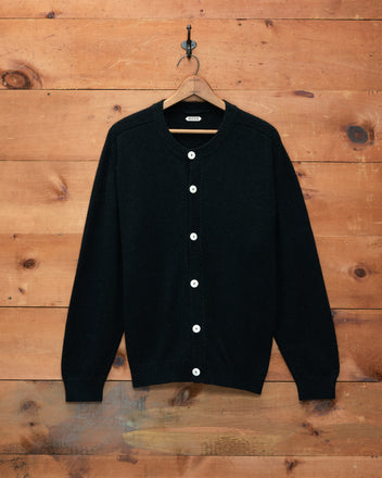 The Orchard Cardigan - Black, displayed on a hanger against a wooden wall, features antique pearl buttons and is crafted from luxuriously soft alpaca merino wool.