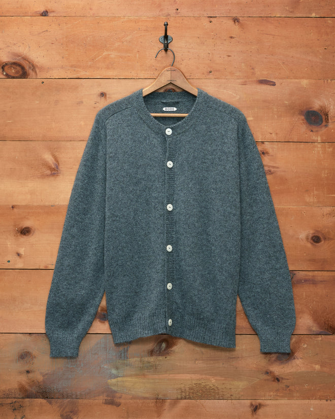 The Orchard Cardigan - Grey hangs on a wooden hanger against a rustic wooden wall, featuring white buttons and crafted from merino wool.
