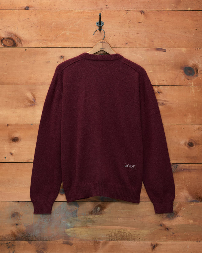 The Orchard Cardigan - Maroon, made from merino wool, hangs on a wooden wall facing backward, with “808C” stitched near the lower right hem.