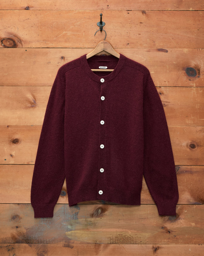 The Orchard Cardigan - Maroon, featuring a classic fit and white buttons, is displayed on a wooden hanger against a wooden wall backdrop.