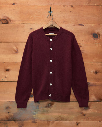 The Orchard Cardigan - Maroon, featuring a classic fit and white buttons, is displayed on a wooden hanger against a wooden wall backdrop.