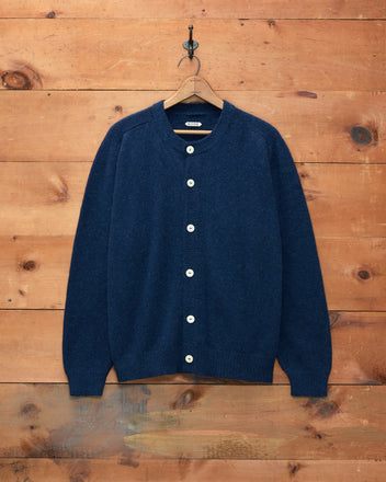 The Orchard Cardigan - Navy, crafted from merino wool and featuring white buttons, hangs on a wooden hanger against a wooden wall.