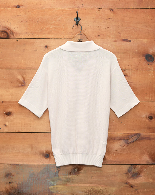 The Boxy Polo in Ecru, a short-sleeved knit shirt with a boxy silhouette, hangs on a hook against a wooden wall and is shown from the back.