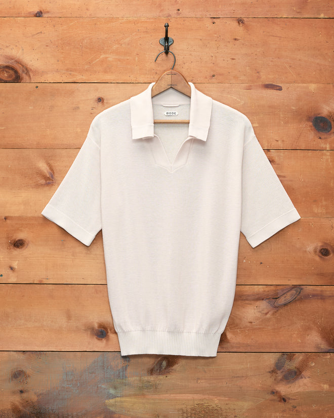 The Boxy Polo - Ecru, a short-sleeve knit shirt with a boxy silhouette in light beige, hangs on a wooden hanger against a wooden wall.