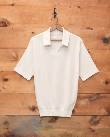 The Boxy Polo - Ecru, a short-sleeve knit shirt with a boxy silhouette in light beige, hangs on a wooden hanger against a wooden wall.