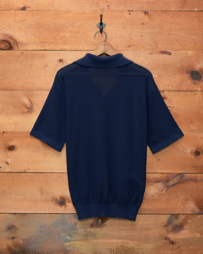 The Boxy Polo - Navy, featuring short sleeves and a boxy silhouette, is displayed from the back as it hangs on a wooden wall.