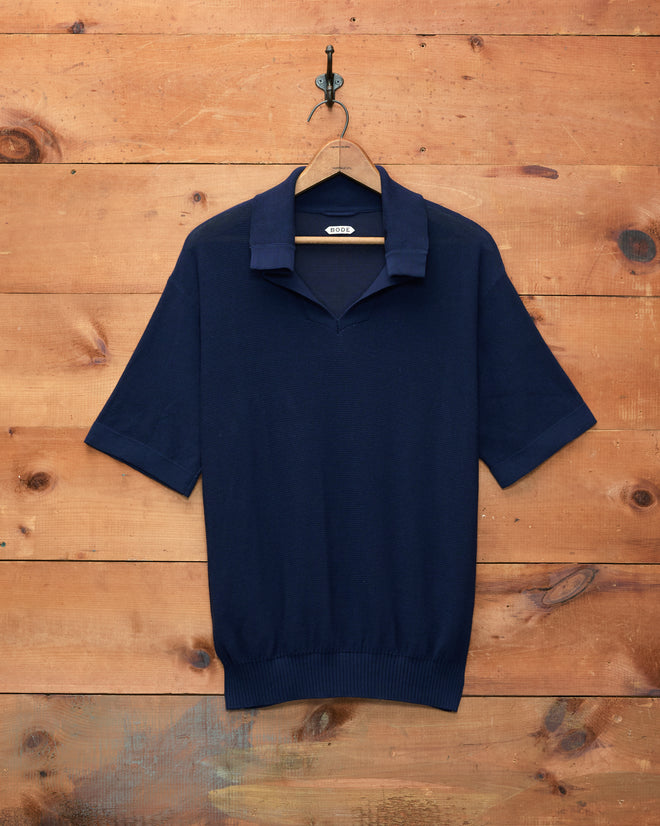 The Boxy Polo - Navy, a short-sleeve knit shirt with a boxy silhouette, is displayed on a black hanger hanging against a wooden wall.