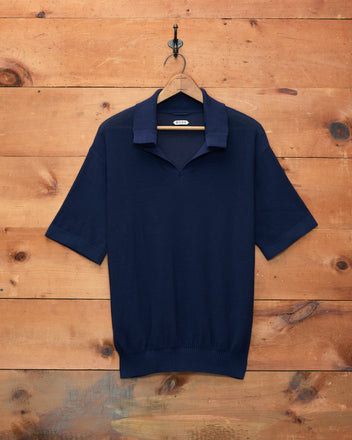 The Boxy Polo - Navy, a short-sleeve knit shirt with a boxy silhouette, is displayed on a black hanger hanging against a wooden wall.