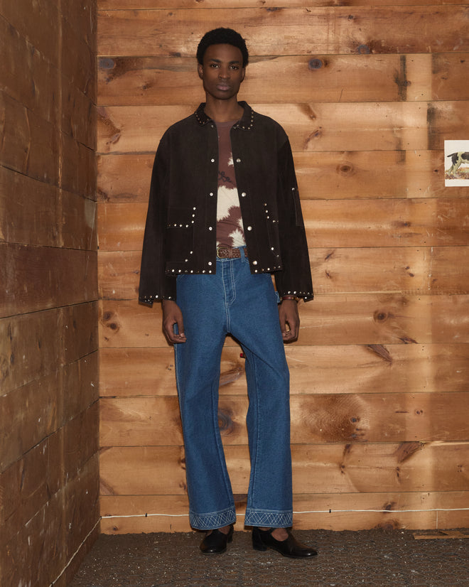 A person wearing a chocolate brown Studded Welding Jacket, tie-dye shirt, blue jeans, and black shoes stands against a wooden wall background.