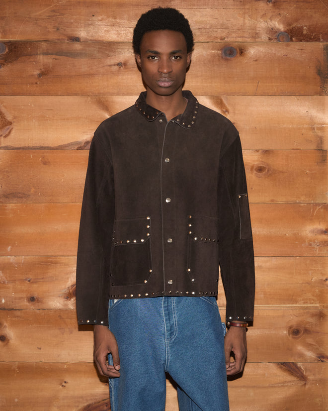 A person in a Studded Welding Jacket - Brown and blue jeans stands against a wooden wall.