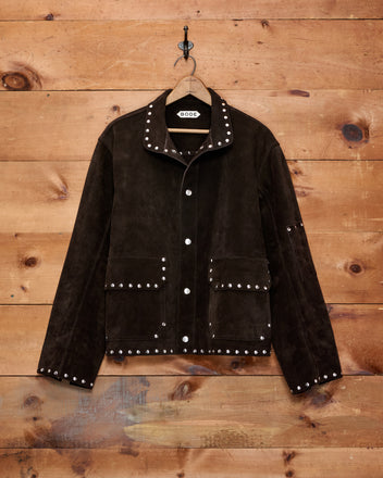 A dark brown suede jacket with silver stud embellishments, hanging on a wooden hanger against a rustic wood plank wall. The jacket features a band collar, snap-button front, and two front flap pockets.