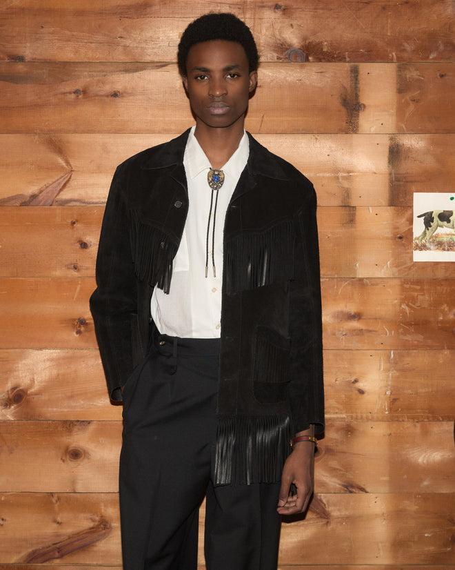 A person in the Appalachian Jacket – Black, featuring hand-cut fringe, styled with a bolo tie and white shirt, stands against a wooden wall.