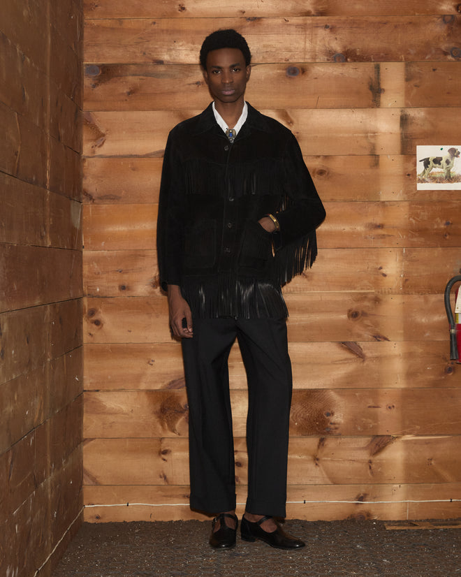 A person in the Appalachian Jacket - Black, featuring hand-cut fringe, and matching pants stands against wooden walls, next to a small art piece and a cart.