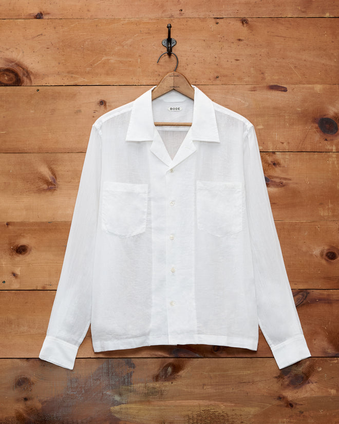 A Voile Long Sleeve Shirt in white, featuring a boxy silhouette and two chest pockets, hangs on a wooden wall.