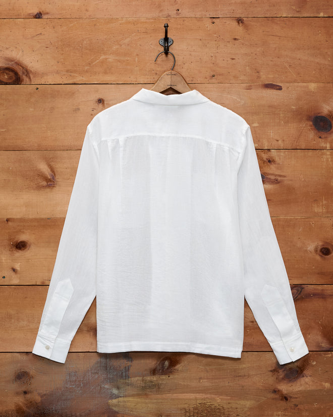 The Voile Long Sleeve Shirt - White, featuring a boxy silhouette, hangs on a hook against a wooden wall and is shown from the back.