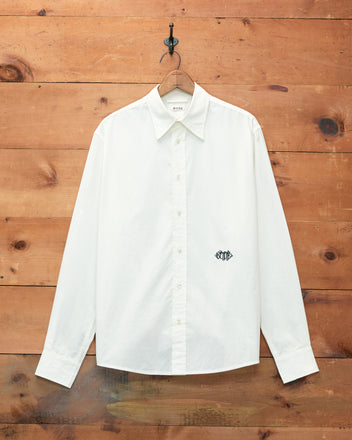 The Monogrammed Poplin Shirt, a white long-sleeve button-down, hangs on a wooden wall and showcases a small black embroidered Bode monogram on the chest.