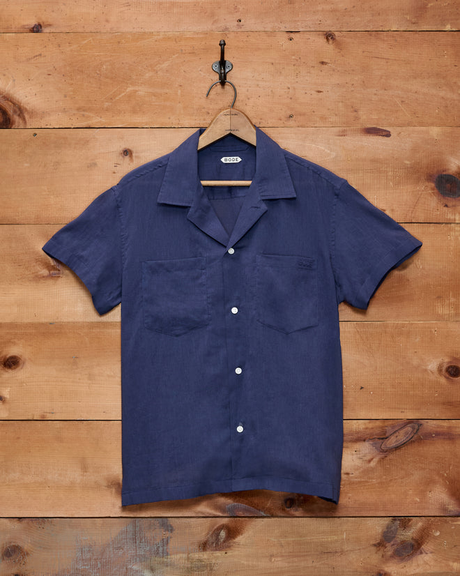 The Voile Short Sleeve Shirt - Navy, featuring a boxy silhouette and crafted from cotton, hangs on a wooden hanger against a wooden wall.
