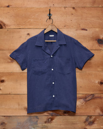 The Voile Short Sleeve Shirt - Navy, featuring a boxy silhouette and crafted from cotton, hangs on a wooden hanger against a wooden wall.