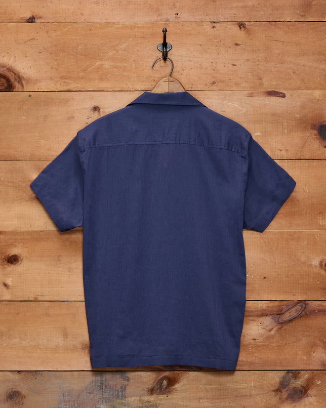 A Voile Short Sleeve Shirt - Navy, featuring a boxy silhouette and crafted from cotton, hangs on a wooden wall and is displayed from the back.