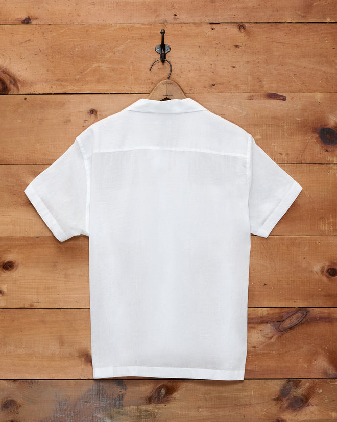 The Voile Short Sleeve Shirt - White hangs on a wooden wall, displayed from the back to highlight its relaxed, boxy silhouette.