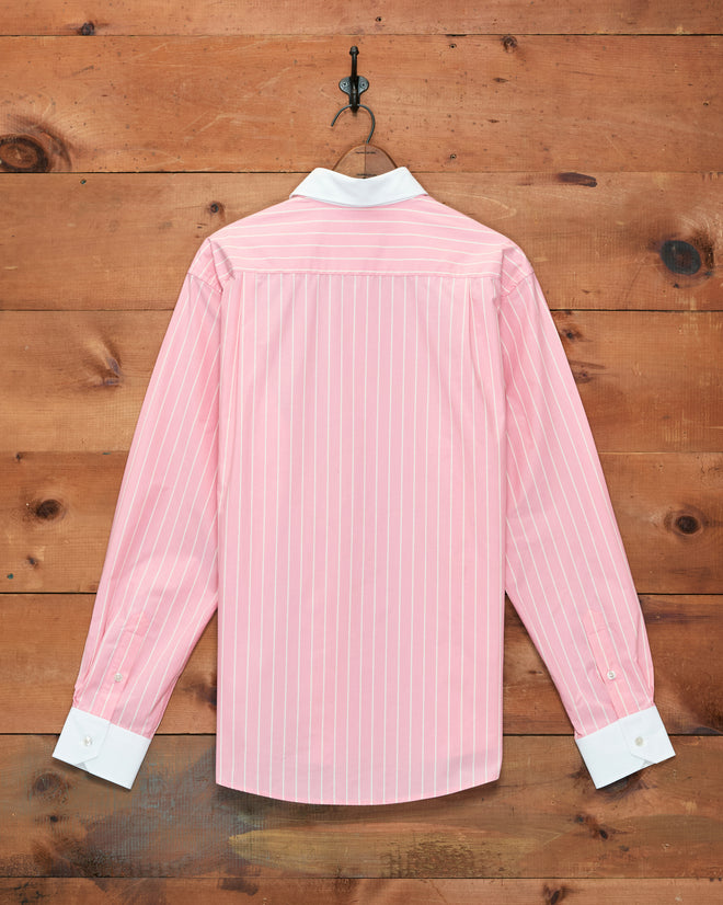 The Pickfair Shirt—a pink pinstripe cotton poplin Winchester shirt with a contrasting white collar and cuffs—hangs on a hook against a wooden wall.