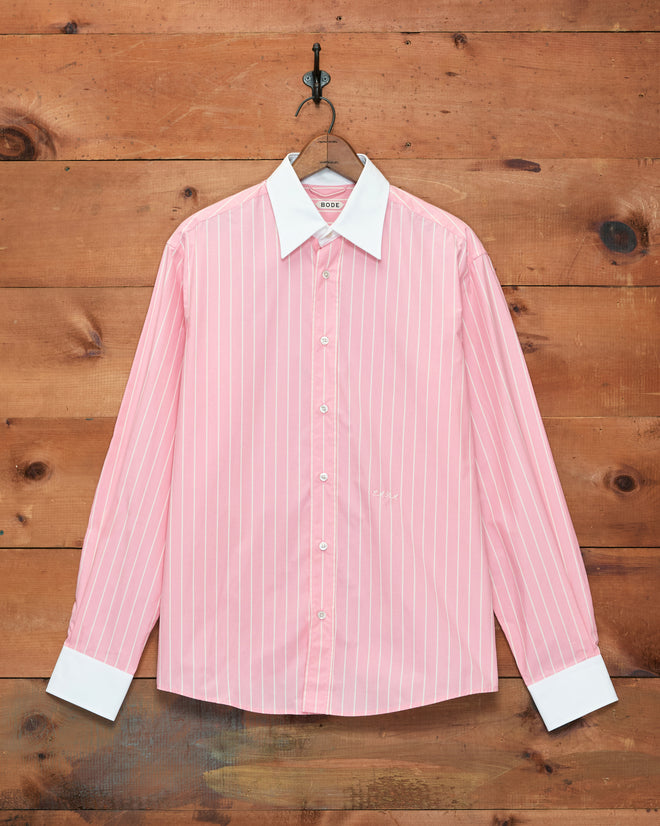 The Pickfair Shirt, a pink cotton poplin dress shirt with white vertical stripes and classic Winchester styling—contrasting white collar and cuffs—is displayed hanging on a wooden wall.